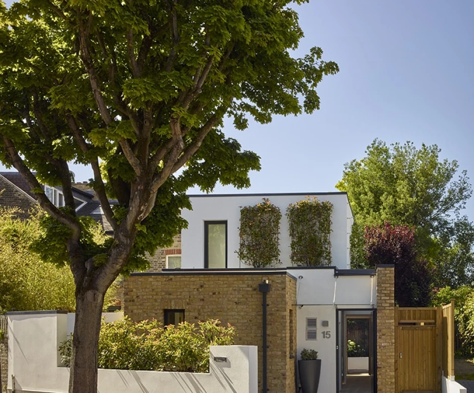 Modern two-story house with a blend of white walls and exposed brick, surrounded by lush greenery. A large tree shades the front, and a wooden fence adds an inviting touch. Bright blue sky above completes the scene.