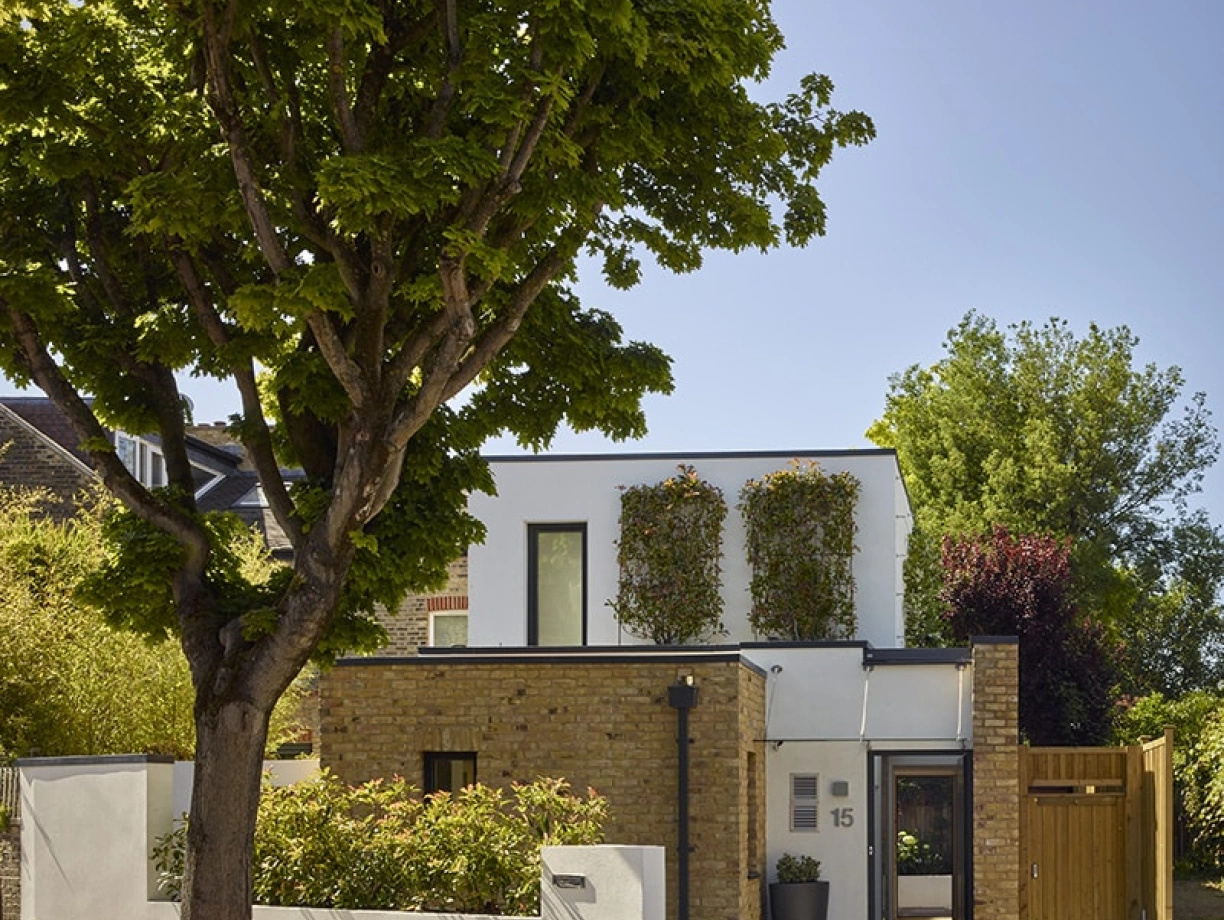 Modern two-story house with a blend of white walls and exposed brick, surrounded by lush greenery. A large tree shades the front, and a wooden fence adds an inviting touch. Bright blue sky above completes the scene.