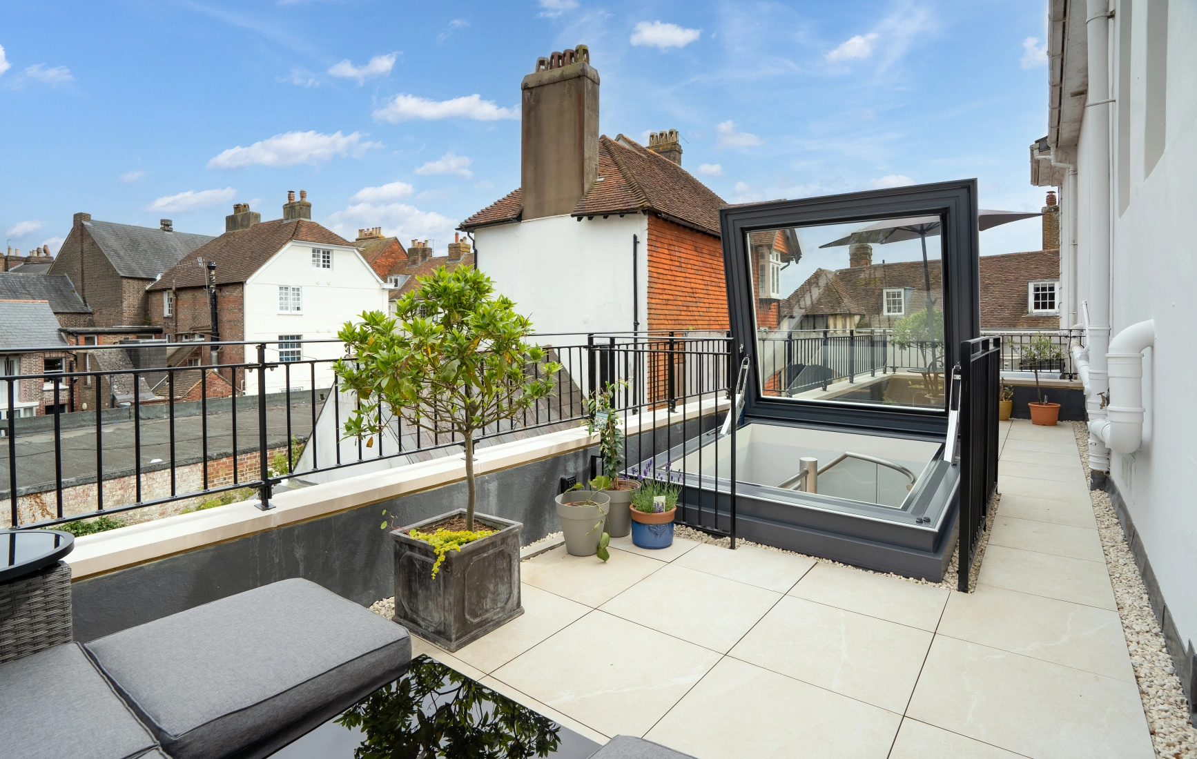 A modern rooftop terrace with a glass skylight, surrounded by a railing. Potted plants add greenery, and a seating area features grey cushions on a black table. Quaint houses are visible in the background under a partly cloudy sky.