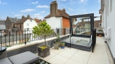 A modern rooftop terrace with a glass skylight, surrounded by a railing. Potted plants add greenery, and a seating area features grey cushions on a black table. Quaint houses are visible in the background under a partly cloudy sky.