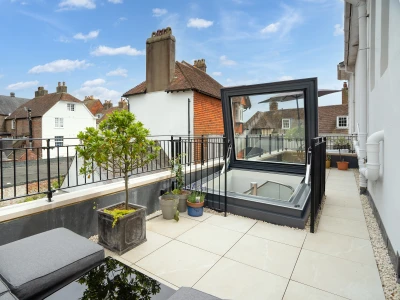 Terrasse moderne avec des carreaux en pierre, un petit arbre dans un pot et un espace lounge. Un puits de lumière est ouvert, offrant une vue sur le ciel bleu parsemé de nuages.