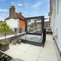 A rooftop terrace featuring modern outdoor furniture, potted plants, and a large skylight that opens up. The background showcases a picturesque view of traditional buildings under a partly cloudy blue sky.