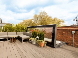 A modern rooftop terrace with gray sectional seating, a small round table, and potted plants. Surrounding greenery adds a natural touch, while a glass windbreak offers a stylish barrier. A blue sky with scattered clouds is visible overhead.