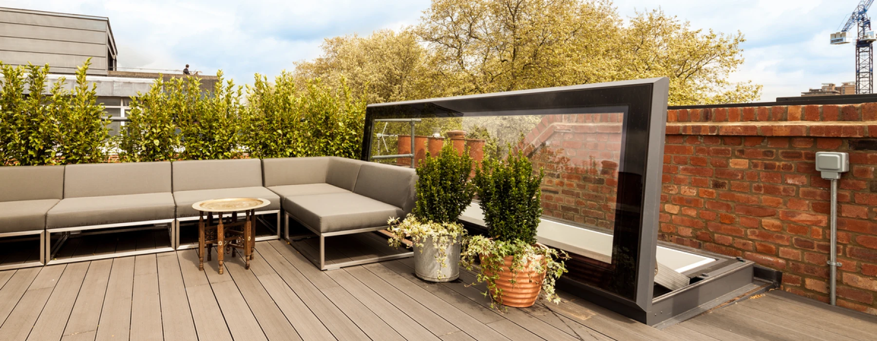 A modern rooftop terrace with gray sectional seating, a small round table, and potted plants. Surrounding greenery adds a natural touch, while a glass windbreak offers a stylish barrier. A blue sky with scattered clouds is visible overhead.