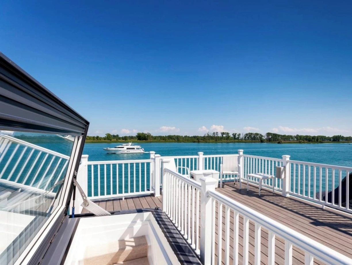 A view from a deck with white railings overlooking a calm blue body of water, featuring a boat cruising in the distance and lush green trees lining the shore under a clear blue sky.