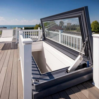 A rooftop with a large hatch opened, revealing a view of the sky and distant water. Two lounge chairs are positioned on the wooden deck, surrounded by a white railing.