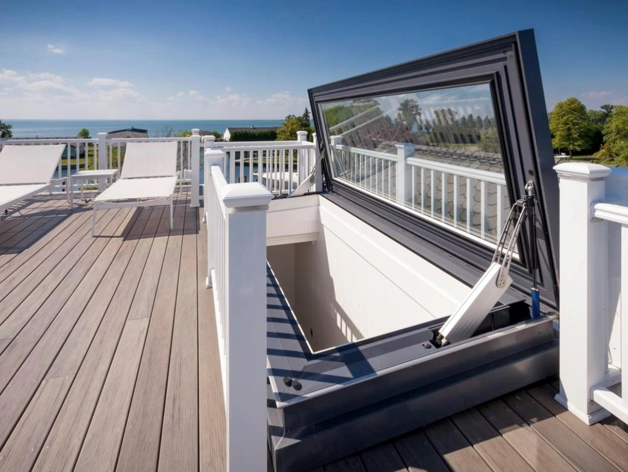 A rooftop deck featuring a large, open hatch that leads below. The surrounding area is adorned with white railings and two lounge chairs, with a scenic view of the sea and blue sky in the background.