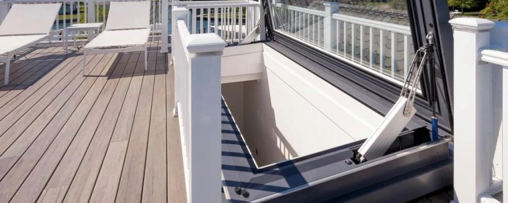 A rooftop with a large hatch opened, revealing a view of the sky and distant water. Two lounge chairs are positioned on the wooden deck, surrounded by a white railing.