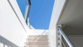 Un escalier blanc avec un tapis beige mène à un espace lumineux sous un ciel bleu. La main courante en métal ajoute une touche moderne.