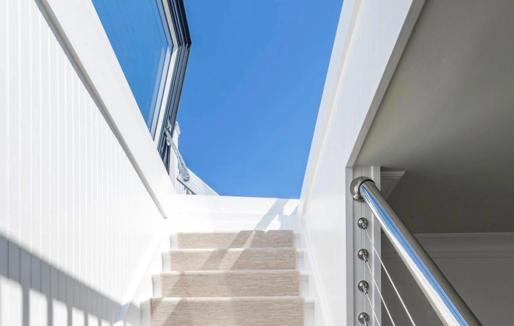 A well-lit staircase with soft beige carpet leading upward, flanked by sleek white walls and a modern railing. The bright blue sky is visible through large windows at the top.