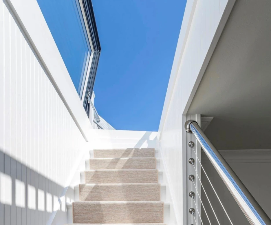 Escalera blanca iluminada por la luz del sol, con un pasamanos de metal y una alfombra beige que cubre los escalones. Al fondo, un cielo azul claro.