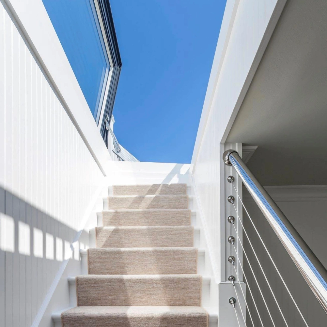 A well-lit staircase with soft beige carpet leading upward, flanked by sleek white walls and a modern railing. The bright blue sky is visible through large windows at the top.