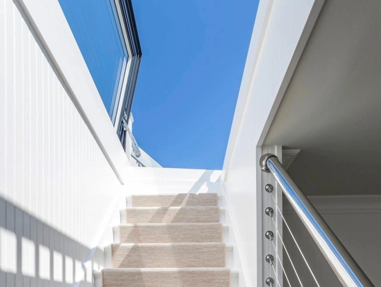 A well-lit staircase with soft beige carpet leading upward, flanked by sleek white walls and a modern railing. The bright blue sky is visible through large windows at the top.