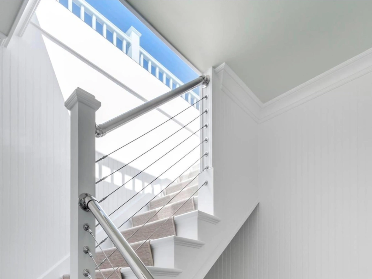 A modern staircase with a sleek metal railing and wooden steps. Natural light streams in from a large window at the top, illuminating the white-paneled walls. A framed picture hangs on the wall near the bottom of the stairs.