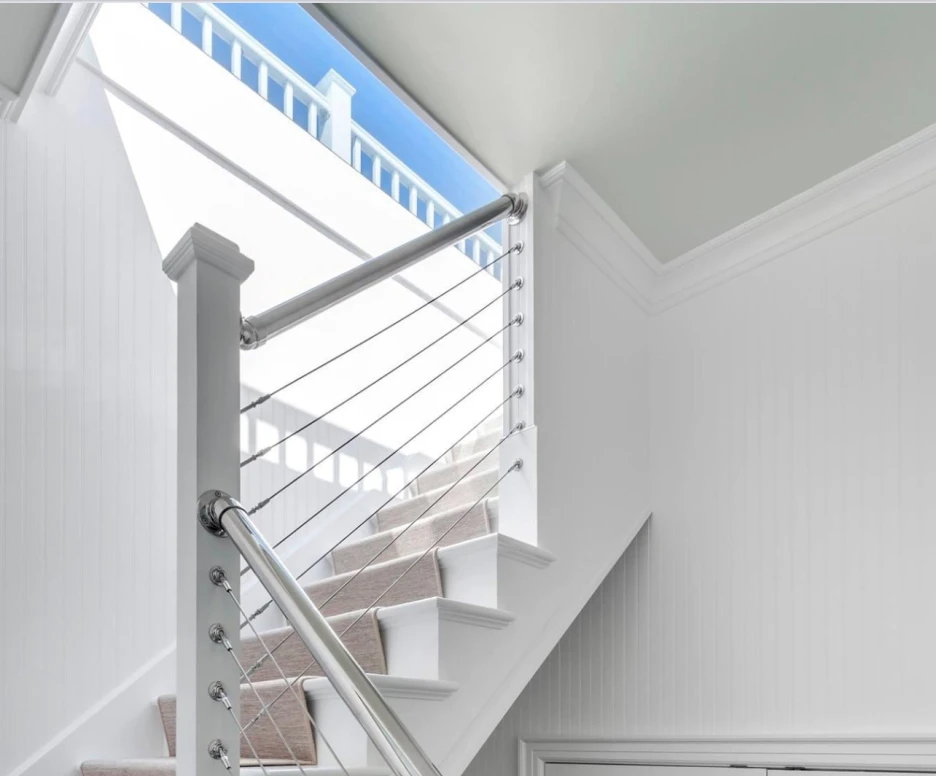 A bright, modern staircase with white walls and a stylish metal railing. Natural light filters through a window at the top, illuminating the carpeted steps. A framed picture hangs on the wall near the base of the stairs.