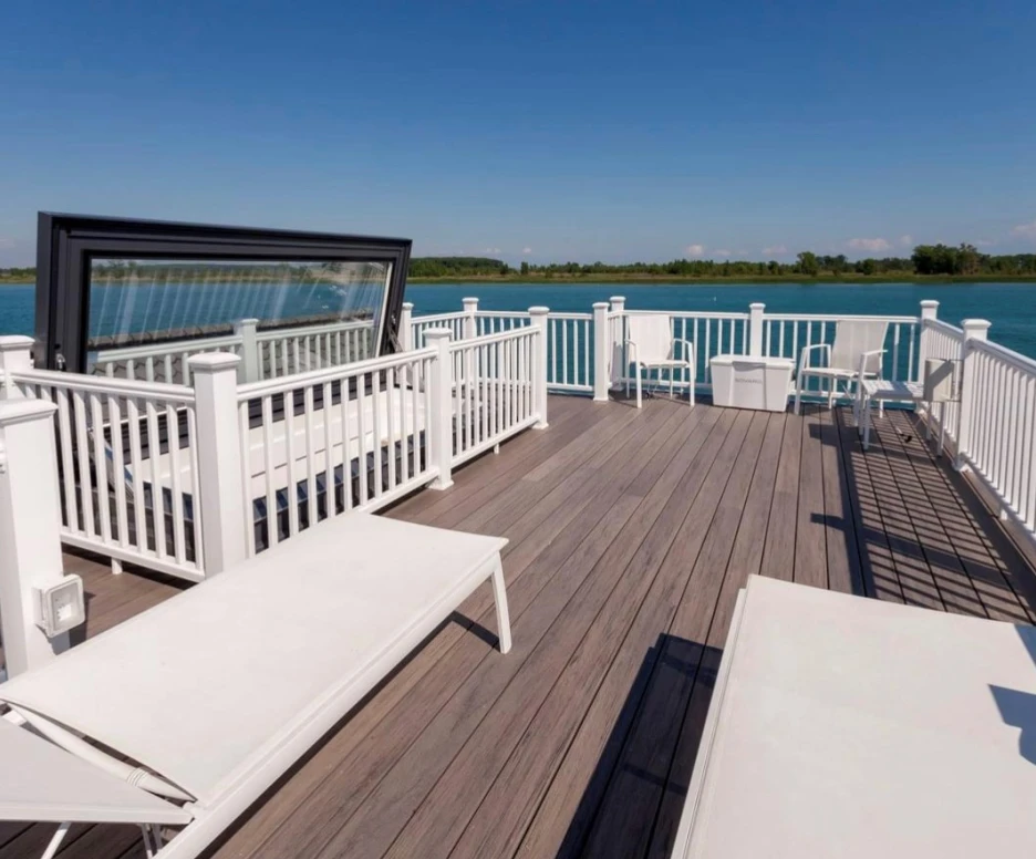 A spacious wooden deck with white lounge chairs and railings overlooks a calm blue body of water, under a clear blue sky.