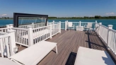 A spacious waterfront deck features white lounge chairs and railings, overlooking a calm blue lake under a clear blue sky.