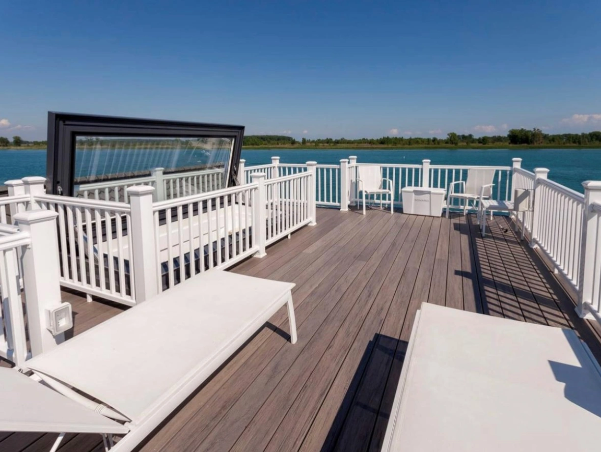 A spacious waterfront deck features white lounge chairs and railings, overlooking a calm blue lake under a clear blue sky.