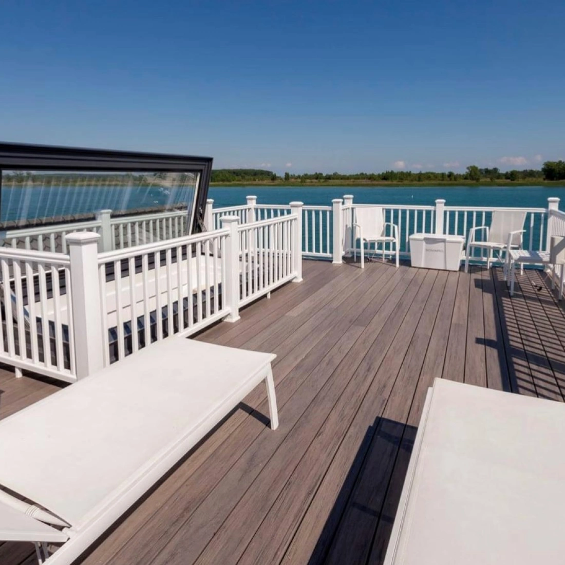 Terrasse moderne en bois avec des chaises longues blanches, offrant une vue sur l'eau calme et le ciel bleu. Des barrières blanches délimitent l'espace.