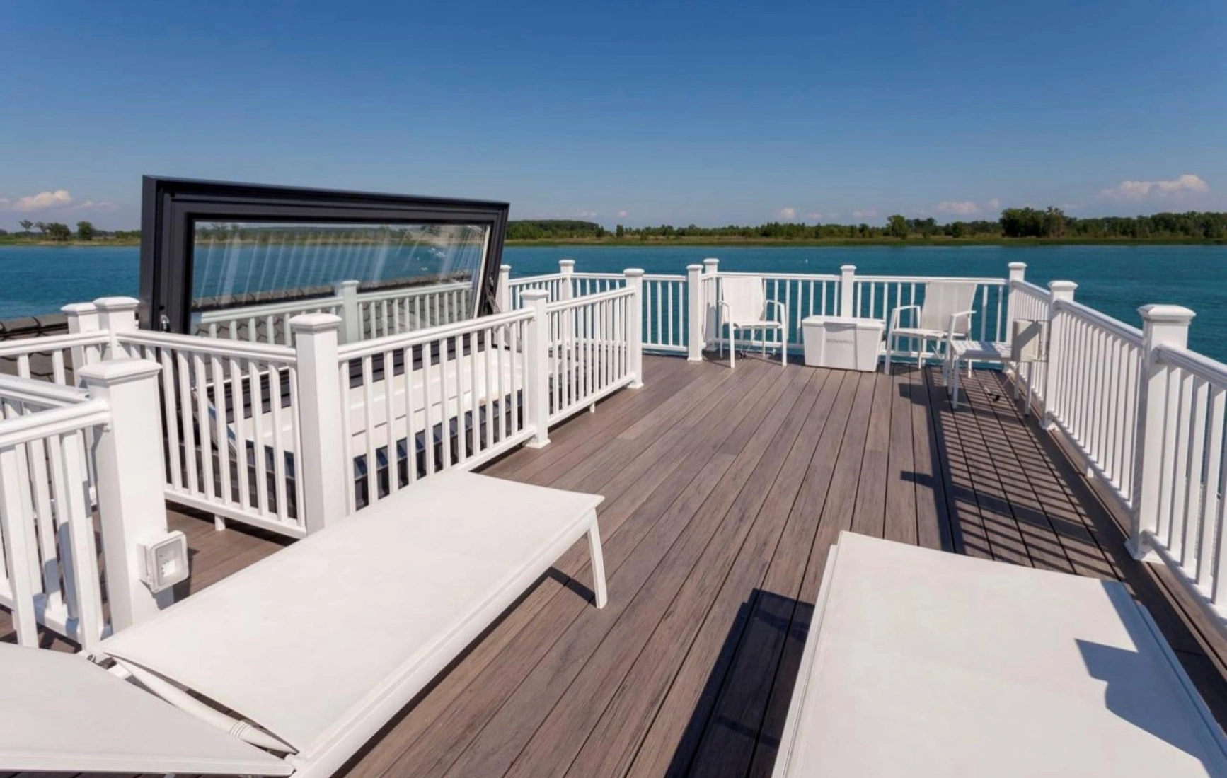 A spacious wooden deck with white lounge chairs and railings overlooks a calm blue body of water, under a clear blue sky.