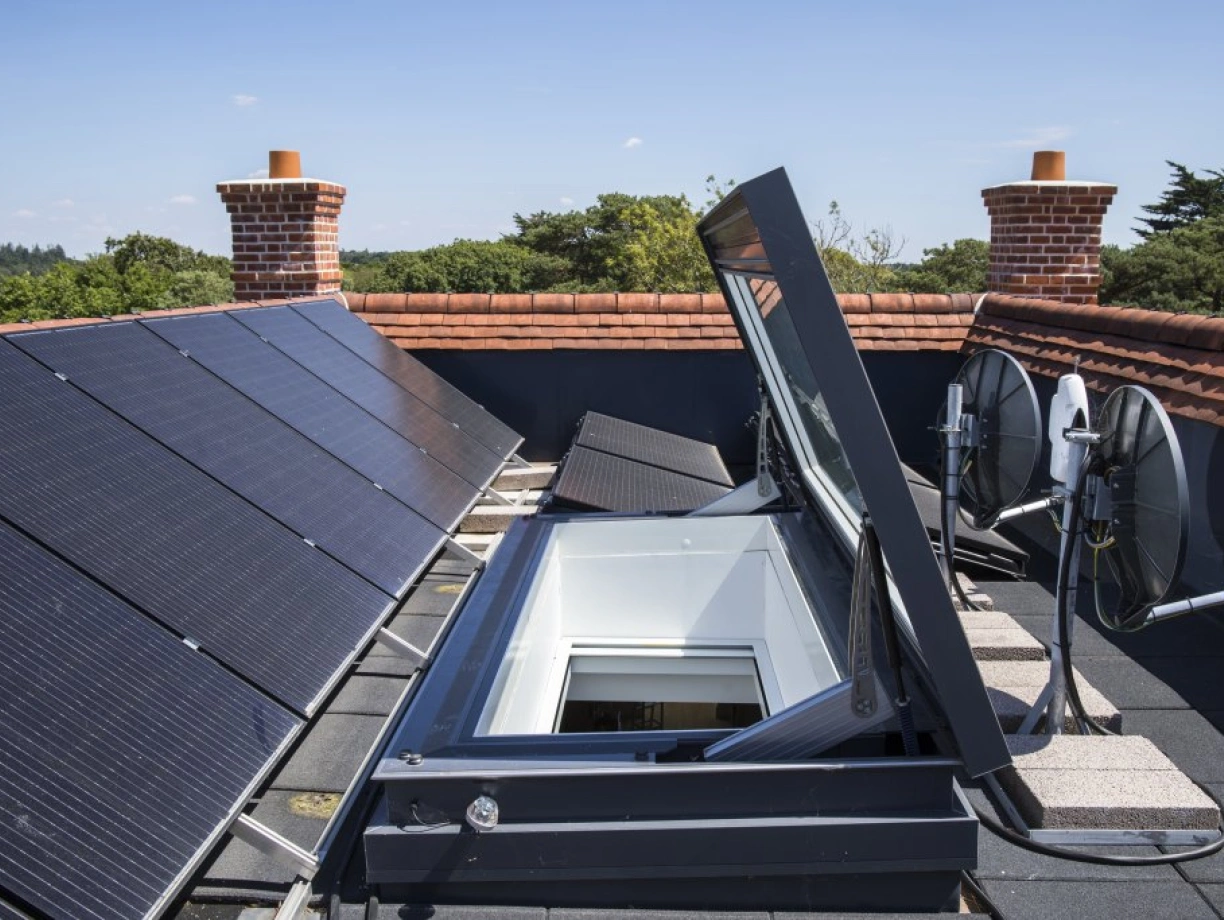 A rooftop featuring solar panels, with a skylight opening and satellite dishes installed nearby. The setting is sunny, with trees visible in the background.