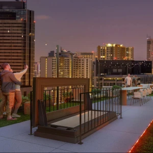 People enjoying a rooftop gathering at dusk, with city skyscrapers illuminated in the background. Some are taking selfies while others are seated at a dining table. String lights add a festive atmosphere to the space.