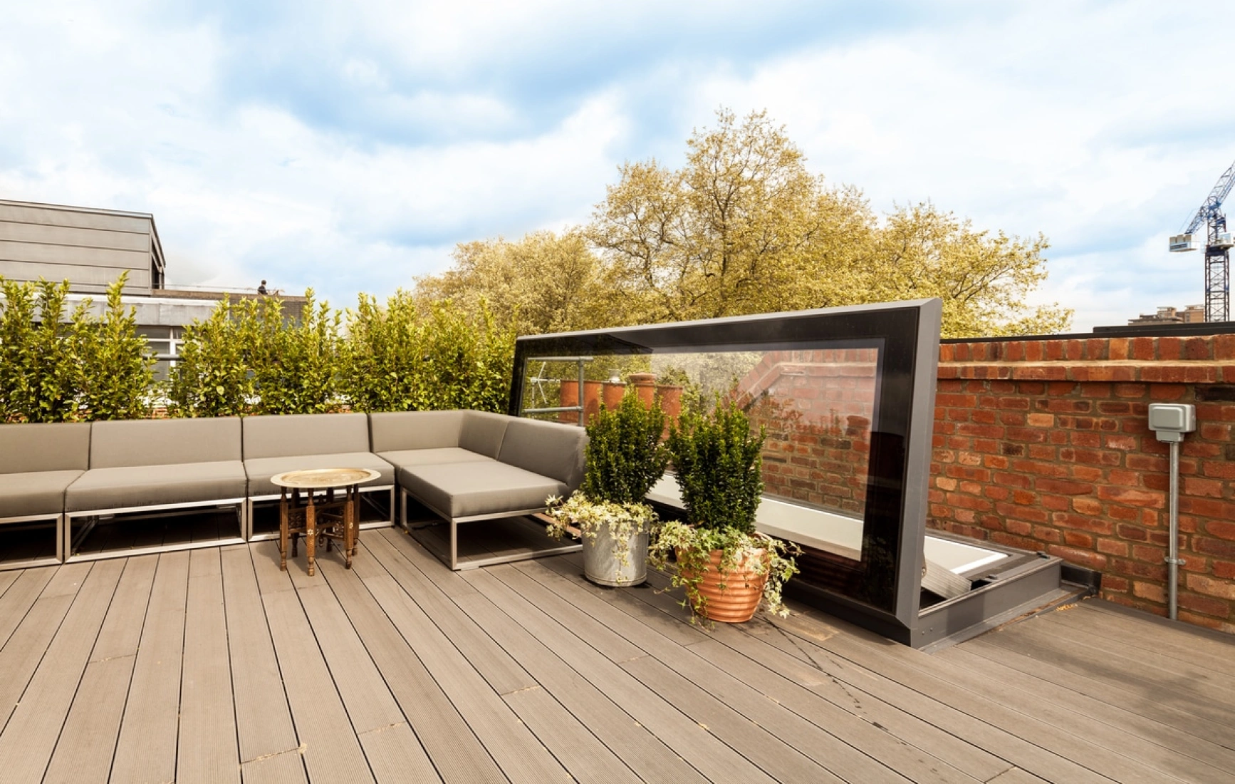 A stylish rooftop patio with gray seating, a round table, and potted plants. The space is surrounded by greenery and features a modern glass partition. Brick walls and a blue sky create a serene atmosphere.