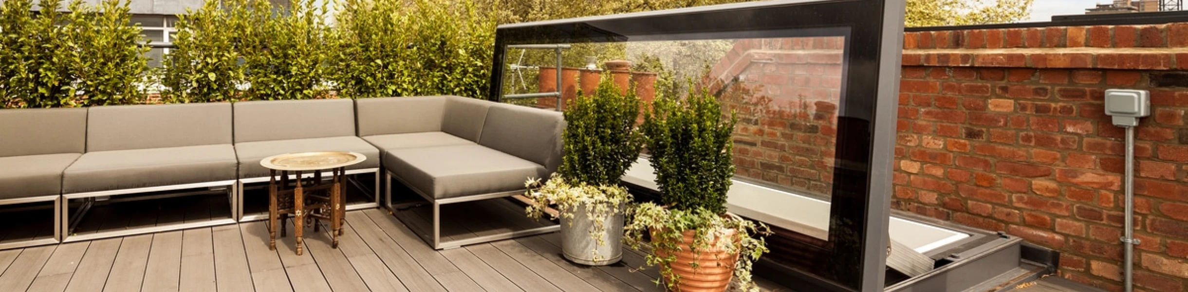 Terraza con muebles de exterior cómodos, incluyendo un sofá, una mesa y plantas en macetas. Al fondo, se aprecian árboles y un cielo despejado.