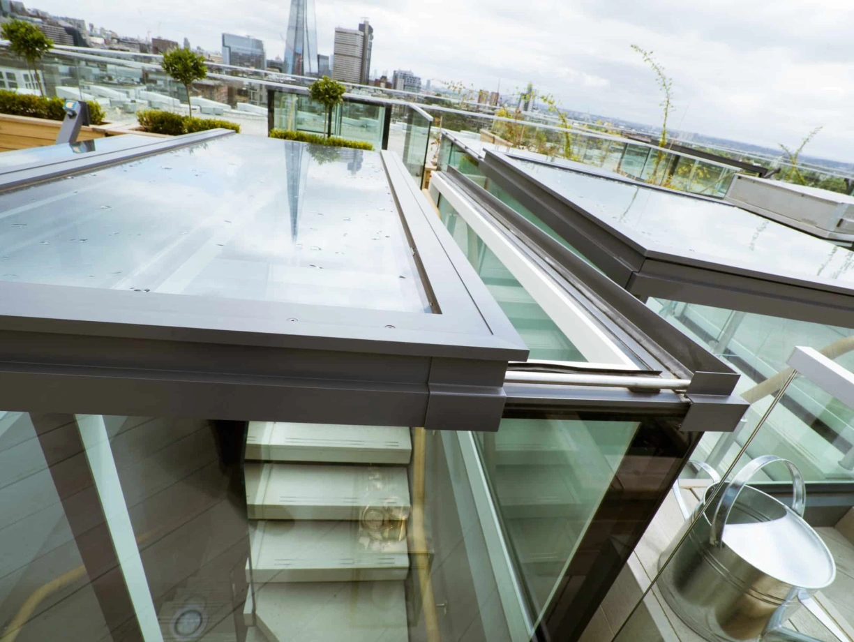 Glass ceiling panels showcase a modern rooftop terrace with views of a city skyline. A staircase is visible below, with metal railings and potted plants. The scene is partly cloudy, hinting at a cool atmosphere.