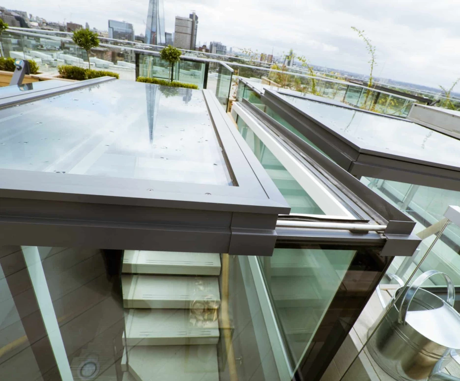 Glass ceiling panels showcase a modern rooftop terrace with views of a city skyline. A staircase is visible below, with metal railings and potted plants. The scene is partly cloudy, hinting at a cool atmosphere.