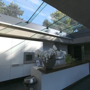 A modern kitchen featuring a sleek design with white cabinetry, a dark countertop, and large glass skylights that allow natural light to flood the space. An arrangement of white orchids adds a decorative touch.
