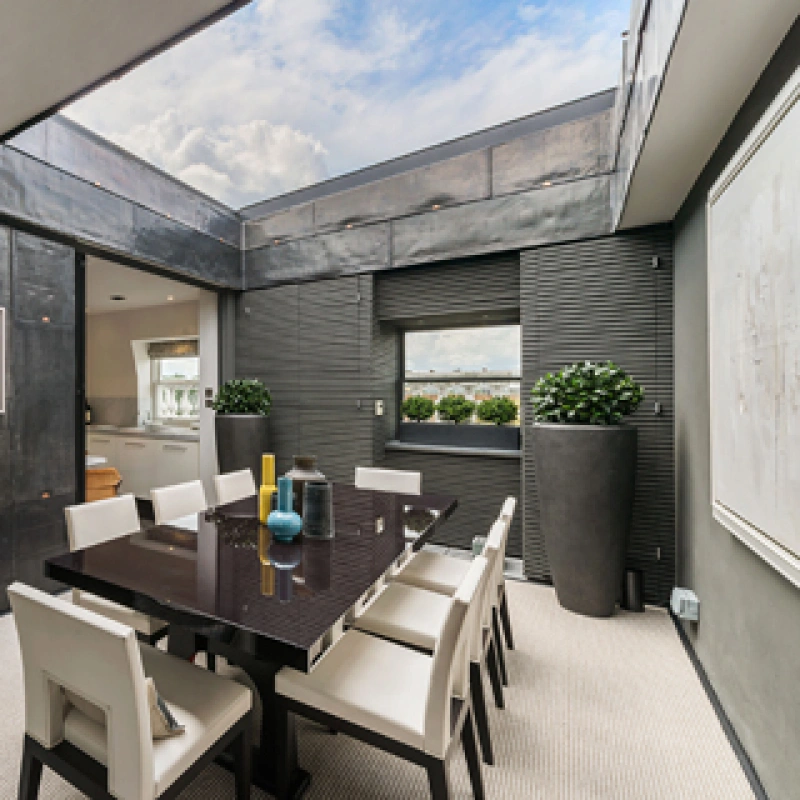 A modern dining room features a sleek black dining table surrounded by white chairs. Large windows and a skylight allow natural light to fill the space, with minimalist decor including plants and contemporary artwork on the walls.