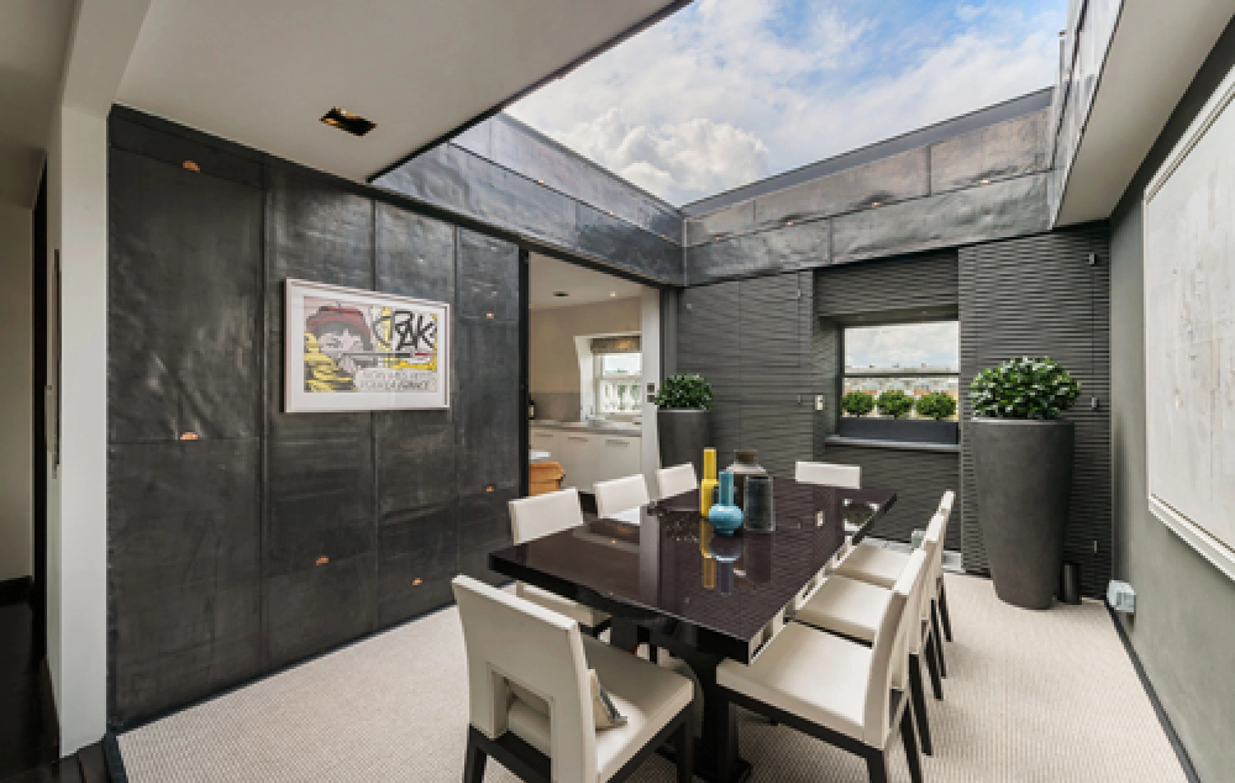 A modern dining room featuring a large dark wooden table surrounded by white chairs. The room has sleek dark walls with a skylight, allowing natural light to enter. Stylish decor includes potted plants and contemporary artwork on the walls.