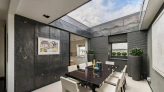 A modern dining room featuring a large dark wooden table surrounded by white chairs. The room has sleek dark walls with a skylight, allowing natural light to enter. Stylish decor includes potted plants and contemporary artwork on the walls.