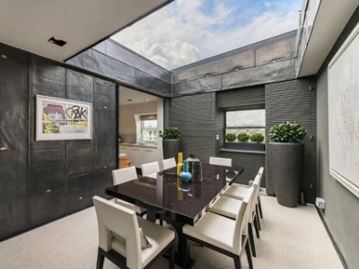 Comedor moderno con una mesa negra rectangular y sillas blancas. Las paredes son de metal oscuro y hay grandes ventanales. Un par de plantas decorativas y una obra de arte en la pared añaden un toque elegante.