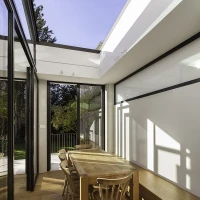 A bright and modern dining area with large glass walls, featuring a wooden table surrounded by four chairs. Sunlight streams in through a skylight and large windows, illuminating the space and offering a view of greenery outside.