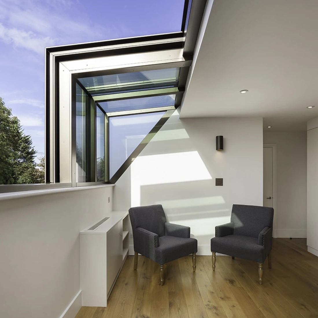 A modern interior with two gray armchairs positioned near a large window, allowing natural light to fill the space. The floor is made of warm wood, and there are minimalist design elements throughout.