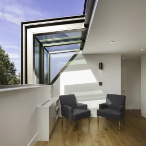 A modern interior with two gray armchairs positioned near a large window, allowing natural light to fill the space. The floor is made of warm wood, and there are minimalist design elements throughout.
