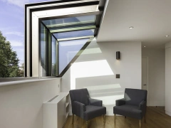 A modern interior with two gray armchairs positioned near a large window, allowing natural light to fill the space. The floor is made of warm wood, and there are minimalist design elements throughout.