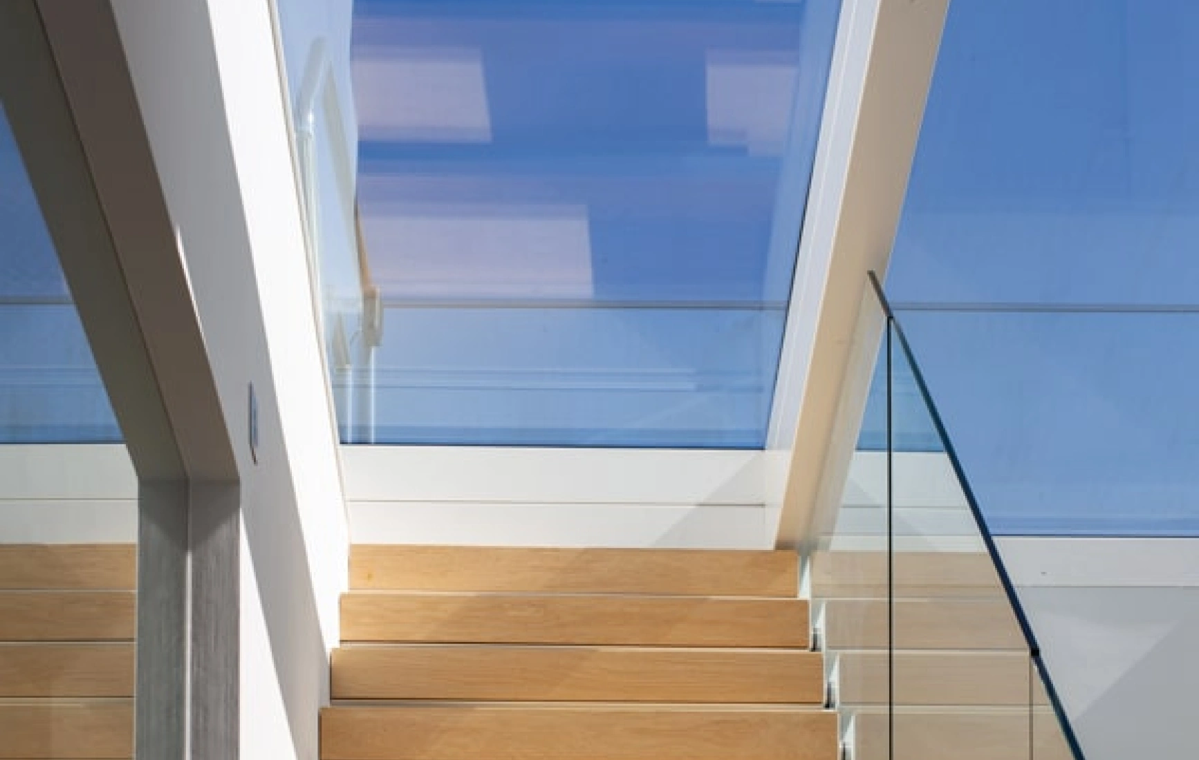 Light-filled staircase with wooden steps leading up to a skylight, featuring glass railings and modern architectural elements.