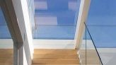 Light-filled staircase with wooden steps leading up to a skylight, featuring glass railings and modern architectural elements.