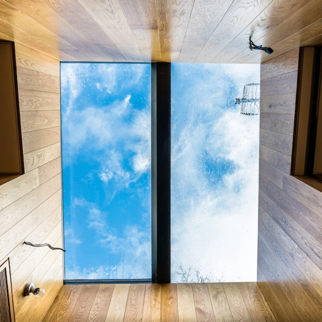 A view from below a skylight showing a clear blue sky with scattered clouds, framed by wooden walls from a modern interior.