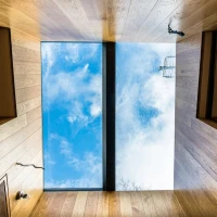 Plafond en bois avec un puits de lumière transparent montrant un ciel bleu avec des nuages, et un reflet d'une structure métallique sur les côtés.