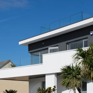 A modern two-story house with a flat roof and a glass railing on the upper balcony. The exterior features a combination of white and gray walls, surrounded by palm trees against a clear blue sky.