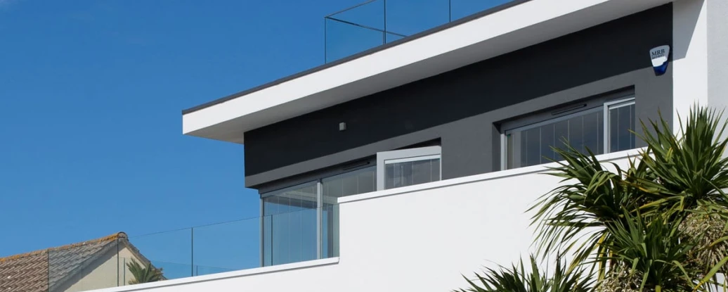 Casa moderna con un diseño minimalista, destacando una terraza con barandilla de vidrio. Jardines tropicales y un cielo despejado de fondo.