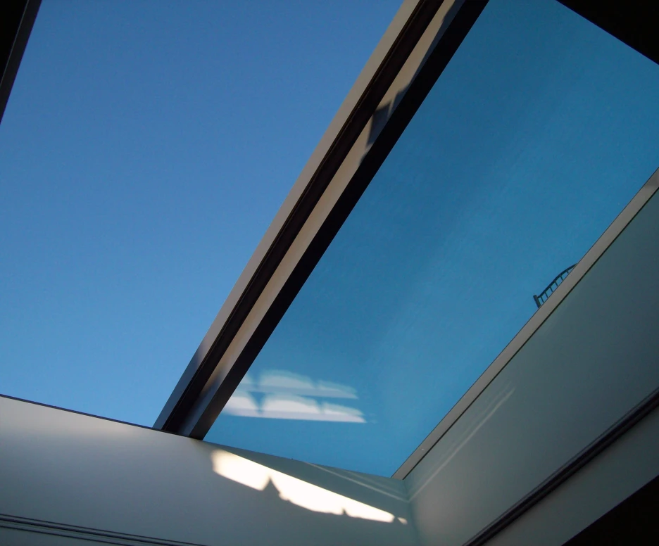 A clear blue sky seen through a modern skylight, with sleek glass framing and a hint of a building silhouette in the background.