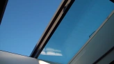 A clear blue sky is visible through a modern skylight, framed by sleek, minimalistic architecture. Light reflects off the glass, creating subtle shadows inside.