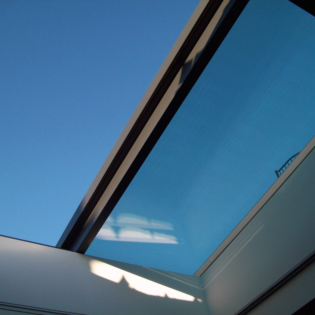 A clear blue sky seen through a modern skylight, with sleek glass framing and a hint of a building silhouette in the background.