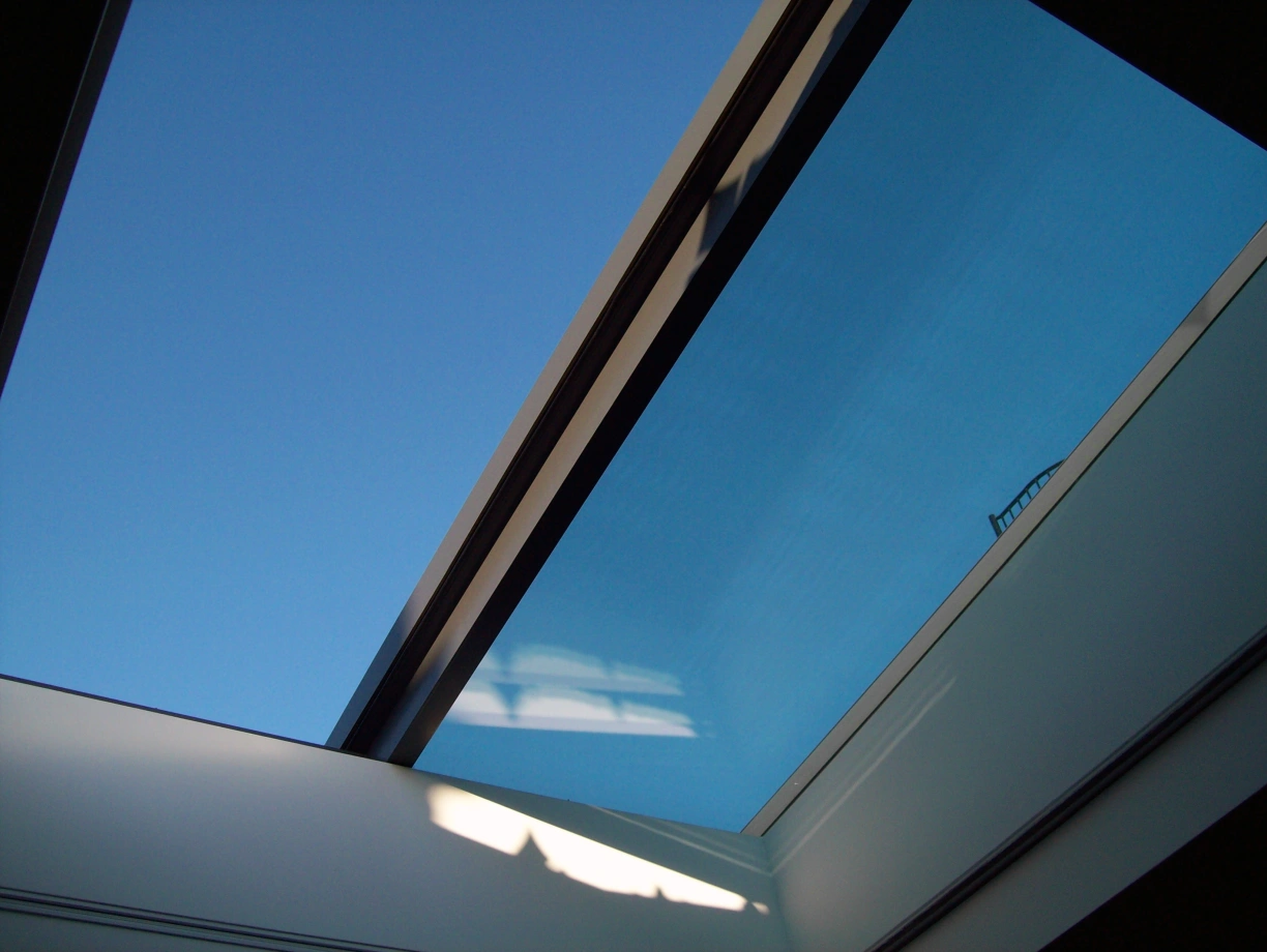 A clear blue sky is visible through a modern skylight, framed by sleek, minimalistic architecture. Light reflects off the glass, creating subtle shadows inside.