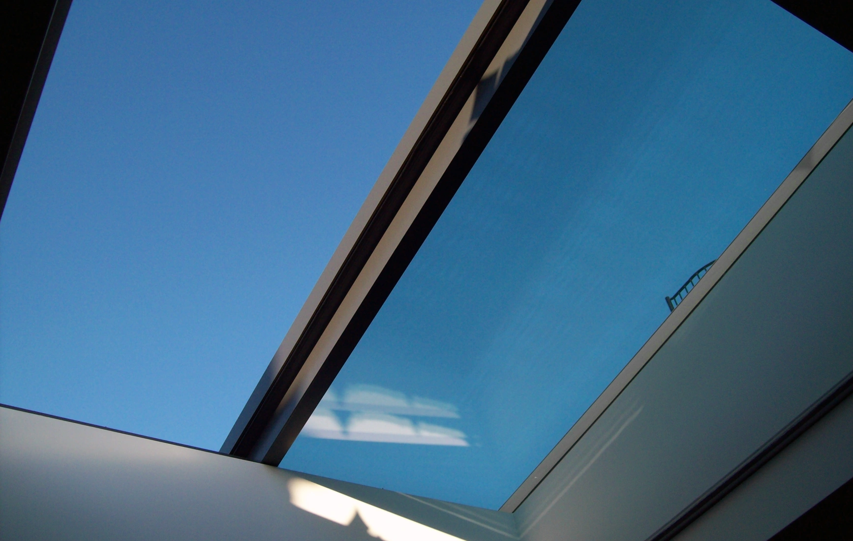 A clear blue sky is visible through a modern skylight, framed by sleek, minimalistic architecture. Light reflects off the glass, creating subtle shadows inside.